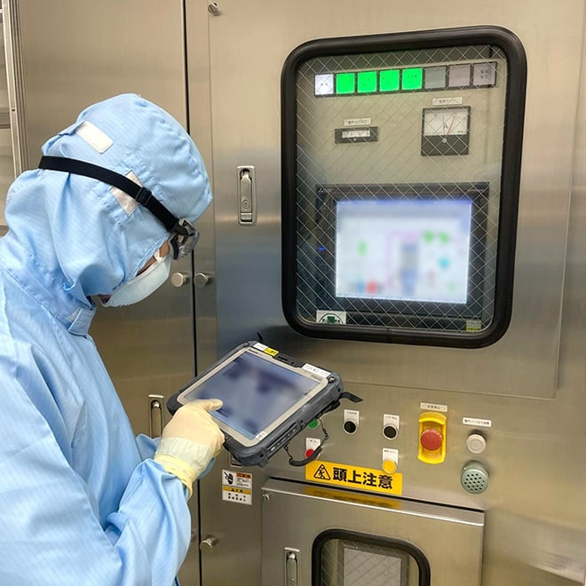 クリーンルーム内で、防護服と手袋、マスクを着用した作業者が、制御盤の前でタブレット端末を操作し、工程ごとの作業状況をリアルタイムで確認している様子