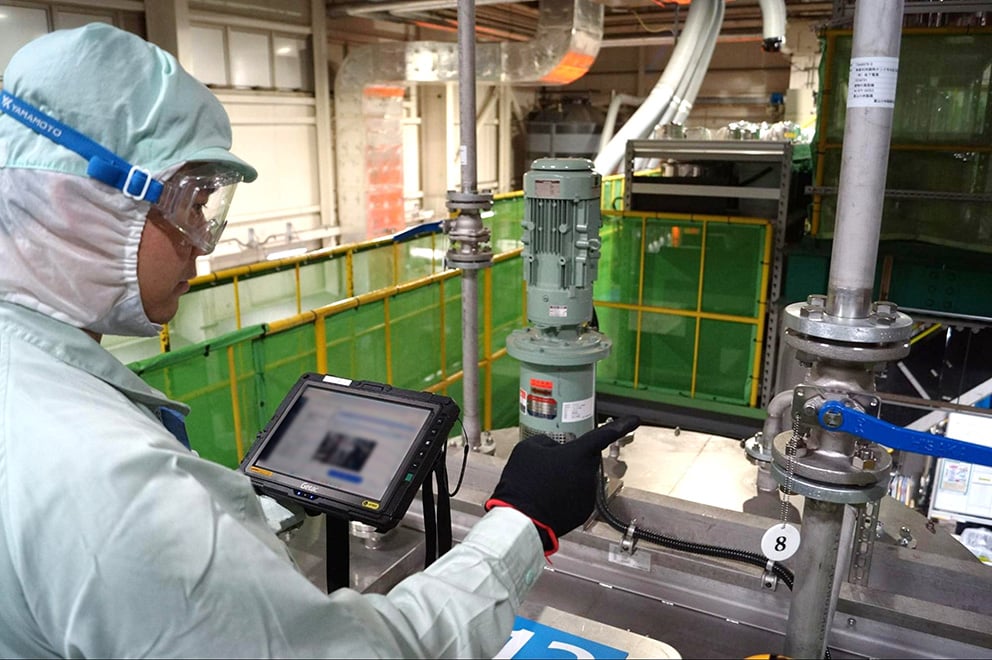 工場内の設備前で、防護服とゴーグル、手袋を着用した作業者がタブレット端末を操作し、配管や装置の状態を確認しながら工程ごとの作業が正常に行われているかをリアルタイムで管理している様子