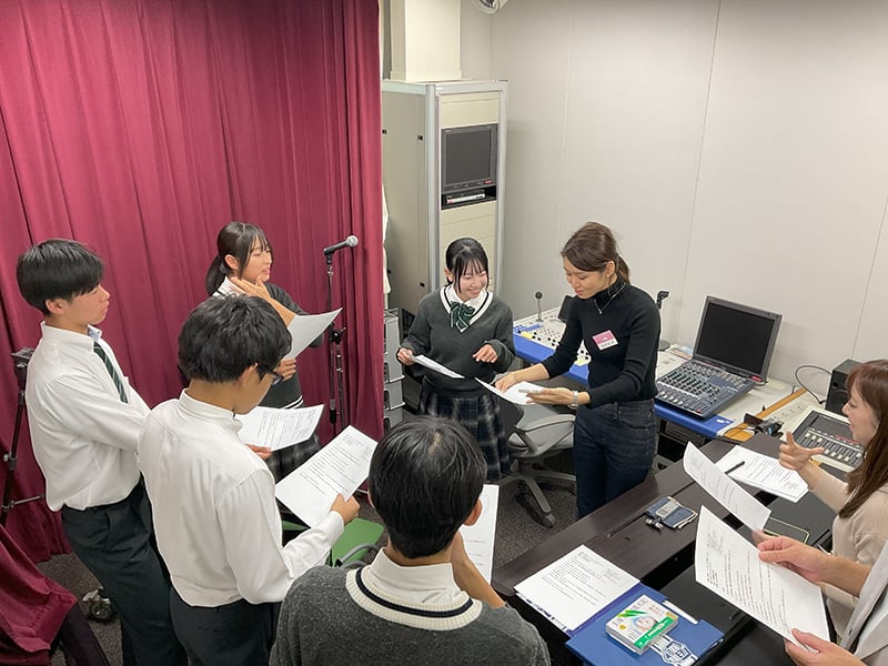 音響設備のある室内で、学生たちが原稿を手に持ち、マイクの前で指導を受けている様子。数名の高校生と、指導役と思われる大人が輪になって原稿を見ながら話し合っており、録音やアナウンスの練習をしている。
