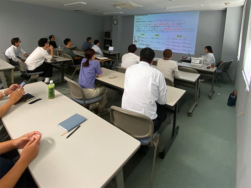 会議室で健康に関するプレゼンテーションを受けている職場の従業員たちの様子。スクリーンには「トリプルリスク」や健康指標に関するスライドが投影されている。