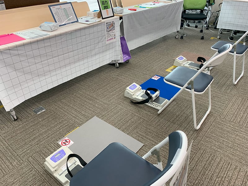 健康イベント会場に設置された体力測定エリア。複数の椅子と足の筋力を測定する装置が並び、奥のテーブルには握力測定器や案内資料も配置されている。
