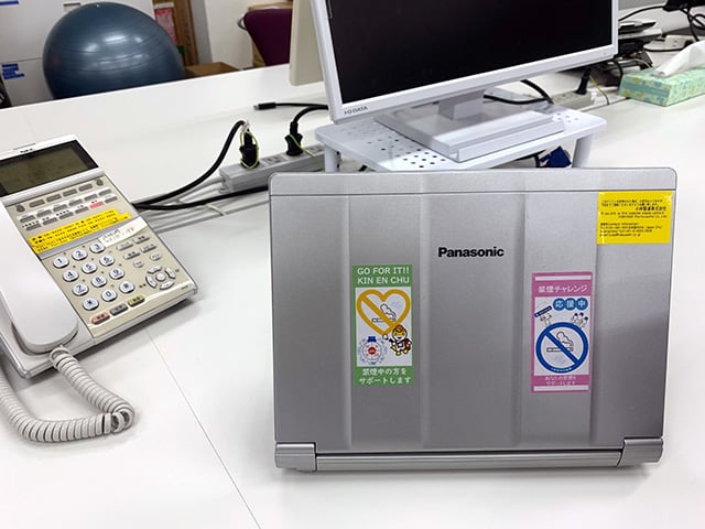 オフィスのデスク上にあるノートパソコンの背面に、禁煙チャレンジと応援メッセージ入りのシールが貼られている様子。隣には電話機やモニターも設置されている。