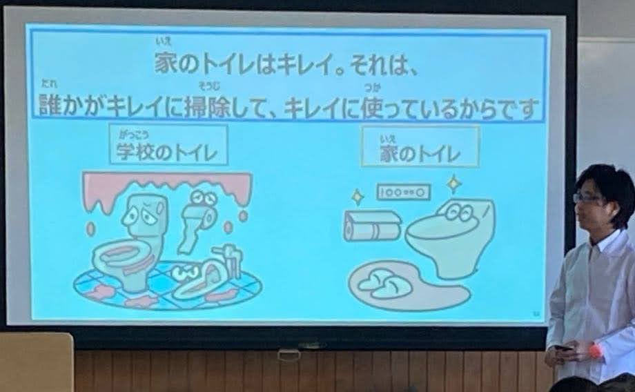 教室内の授業風景で、スクリーンには「家のトイレはキレイ。それは、誰かがキレイに掃除して、キレイに使っているからです」と書かれている。左には汚れた「学校のトイレ」、右には清潔で笑顔の「家のトイレ」のイラストが対比的に描かれており、教師がその前で立って説明している様子。