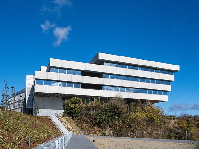 青空の下、横長の窓が並ぶ白い現代的な建物が、植栽のある丘の上に建っている